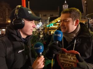 Thorsten Kornblum im Interview mit Lennart Wenk, beide halten ein Mikrofon in der Hand und unterhalten sich. Thorsten Kornblum trägt ein Lebkuchenherz mit der Aufschrift "Gruß vom Braunschweiger Weihnachtsmarkt"