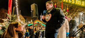 Zu sehen ist eine Weihnachtsmarkt-Kulisse. Ein Radiointerview findet statt. Thorsten Kornblum wird von Lilian Schröder interviewt. Beide halten ein Mikron in der Hand.