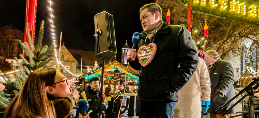 Zu sehen ist eine Weihnachtsmarkt-Kulisse. Ein Radiointerview findet statt. Thorsten Kornblum wird von Lilian Schröder interviewt. Beide halten ein Mikron in der Hand.